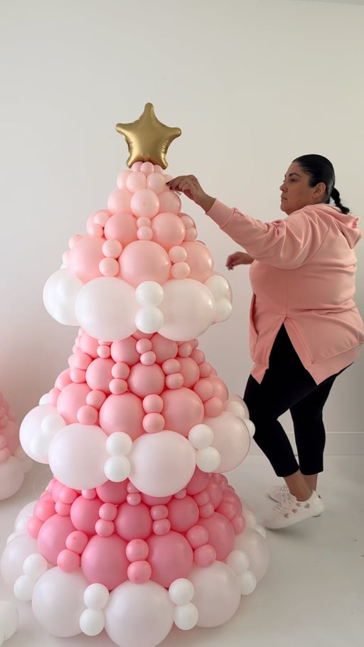 Lilly adjusts the top section of a pink balloon Christmas tree built with layered pink and white balloons and topped with a gold star. She stands beside the structure in a pink hoodie and white sneakers while styling the details. This behind the scenes moment shows the same design taught step by step inside the Whimsical Balloon Christmas Tree Class for holiday installs.