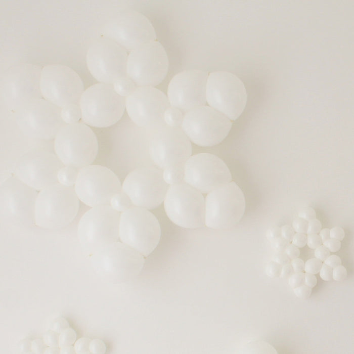 Clustered white balloon snowflakes in multiple sizes lay against a clean white background. Each snowflake is made from small round balloons arranged into layered six-point shapes, creating a bright winter look. The mix of large and small designs adds dimension, making these balloon snowflakes perfect for holiday parties, winter wonderland themes, and seasonal photo backdrops.