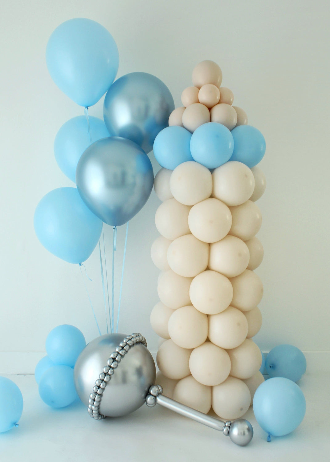 Freestanding baby bottle balloon column paired with a silver baby rattle balloon prop and blue helium balloons, styled for baby showers, newborn welcome decor, and delivery friendly balloon displays. This baby themed balloon setup features neutral and blue tones and showcases a coordinated column and prop design that works for events, hospital pickups, and grab and go balloon orders.