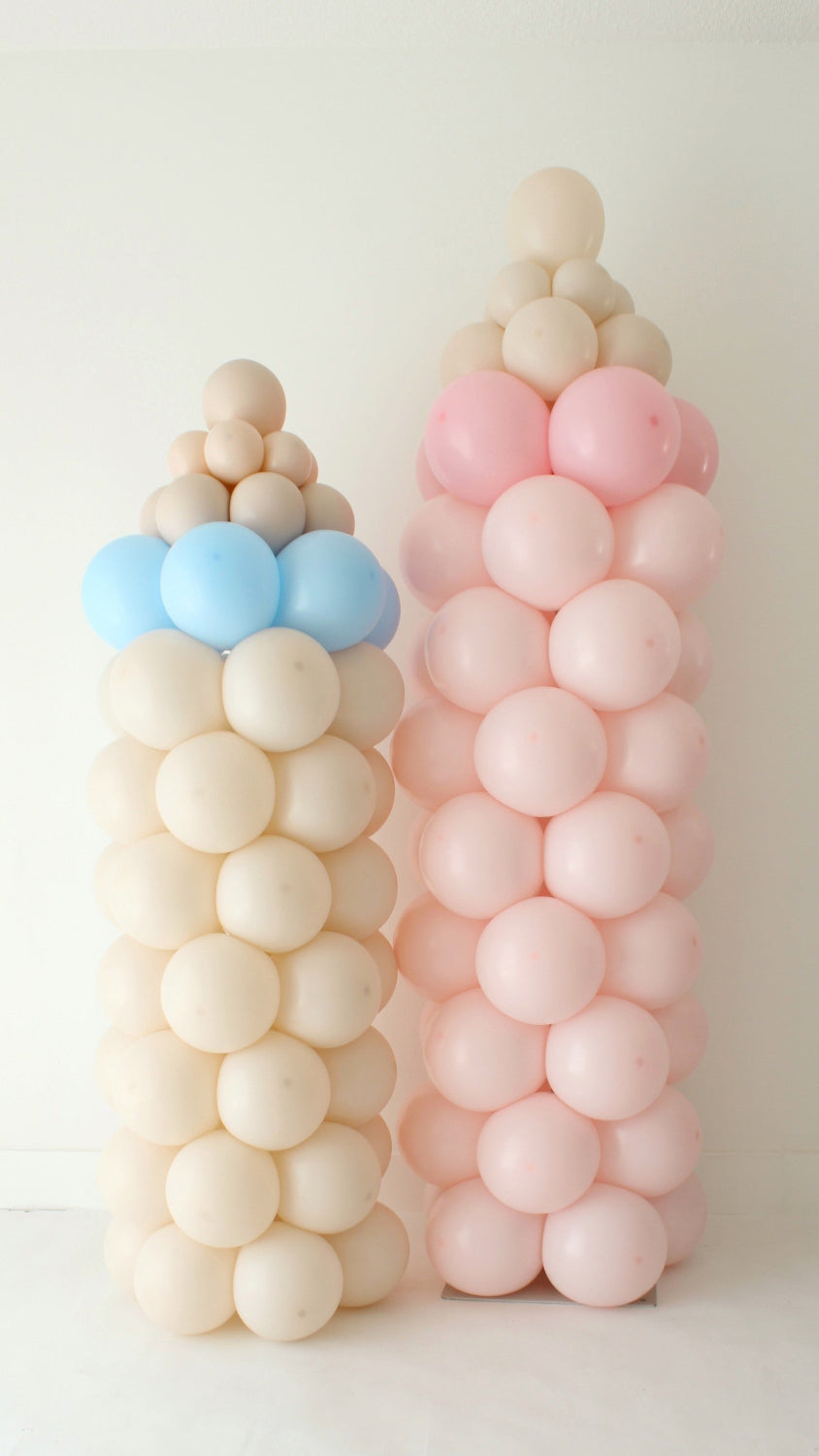 Two baby bottle balloon columns made with pastel pink, blue, and cream balloons, styled as baby shower balloon decor. These freestanding balloon columns are ideal for baby showers, gender reveals, sip and see parties, and welcome baby events. This balloon structure technique can be adapted for different color palettes and themed celebrations.