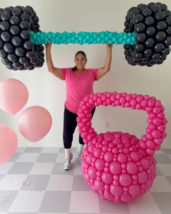 Lilly Jimenez lifting oversized balloon barbell above her head with large pink balloon kettlebell in front, fitness themed balloon decor for gym events, workout parties, and health brand activations.