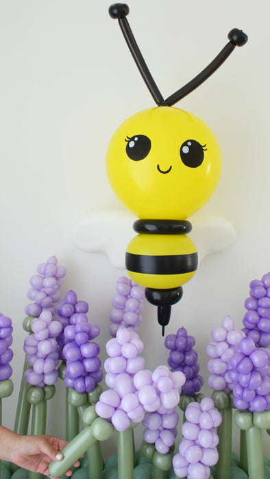 Classic balloon bee sculpture with yellow body, black stripes and white wings floating above purple balloon flowers. Spring garden theme balloon decor designed for birthday parties, baby showers and professional event installations.