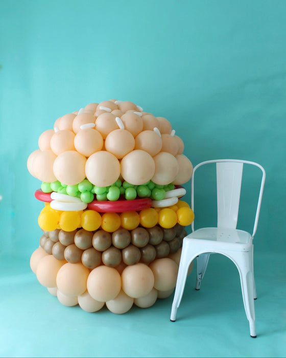 Large balloon burger sculpture next to white chair showing scale on blue background