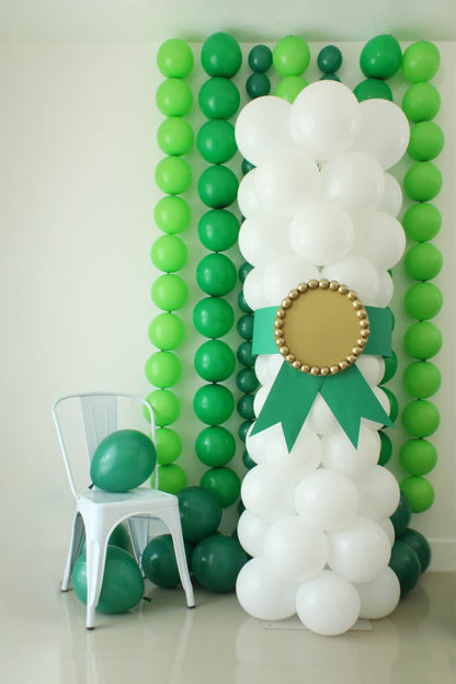 Graduation balloon column styled as a diploma with white balloons, green ribbon, and gold medallion, displayed next to a white chair and layered green balloon backdrop. Perfect for DIY graduation photo booths or stage decorations.