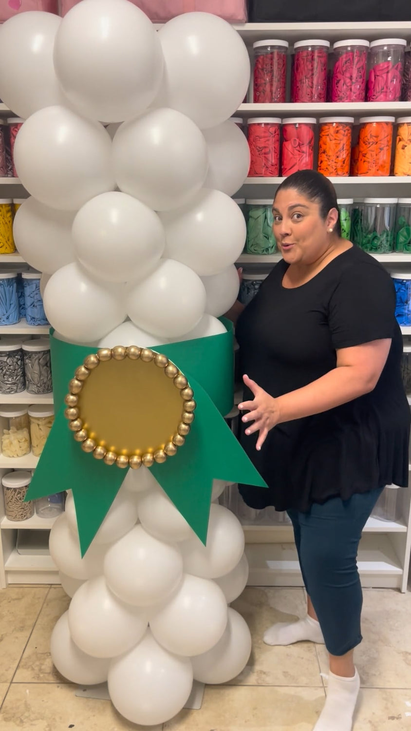 Balloon artist, Lilly Jimenez, standing next to a tall white balloon column designed to look like a graduation diploma, featuring a green ribbon and gold medallion. Perfect for graduations of all ages, this column is part of the DIY Graduation Balloon Diploma Class.
