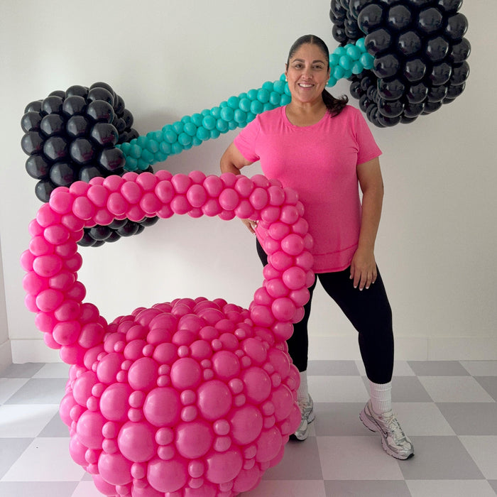 Lilly Jimenez standing next to oversized pink balloon kettlebell with black and teal balloon barbell sculpture, fitness themed balloon decor for gym events, health brand activations, and workout inspired party setups.