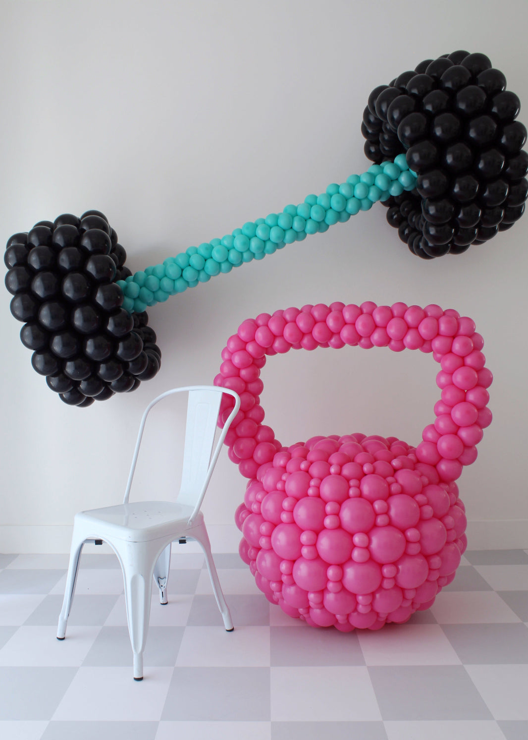 Oversized pink balloon kettlebell and black balloon barbell with teal handle next to white chair, fitness themed balloon decor for gym events, workout parties, and health brand activations.