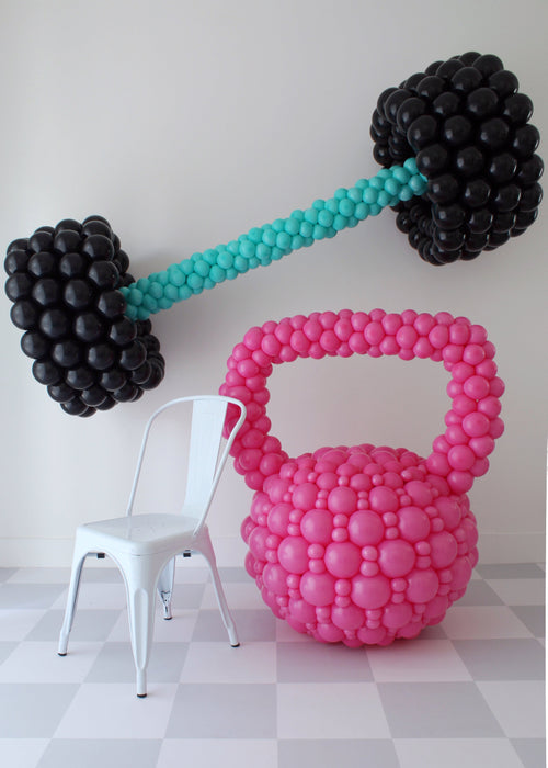 Oversized pink balloon kettlebell and black balloon barbell with teal handle next to white chair, fitness themed balloon decor for gym events, workout parties, and health brand activations.