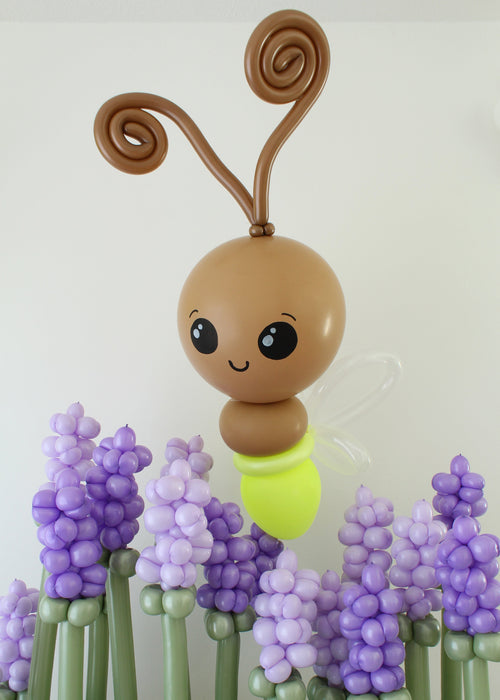 Whimsical glowing light bug/firefly balloon sculpture with brown head, curled antennae and translucent wings displayed among purple balloon flowers. Creative spring balloon decor perfect for garden themes, first birthdays and custom party installations.