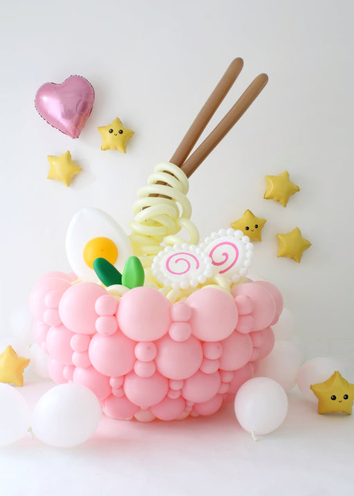 Balloon ramen bowl centerpiece with pink balloon base, spiral noodles, egg, fish cake, and floating chopsticks on white background