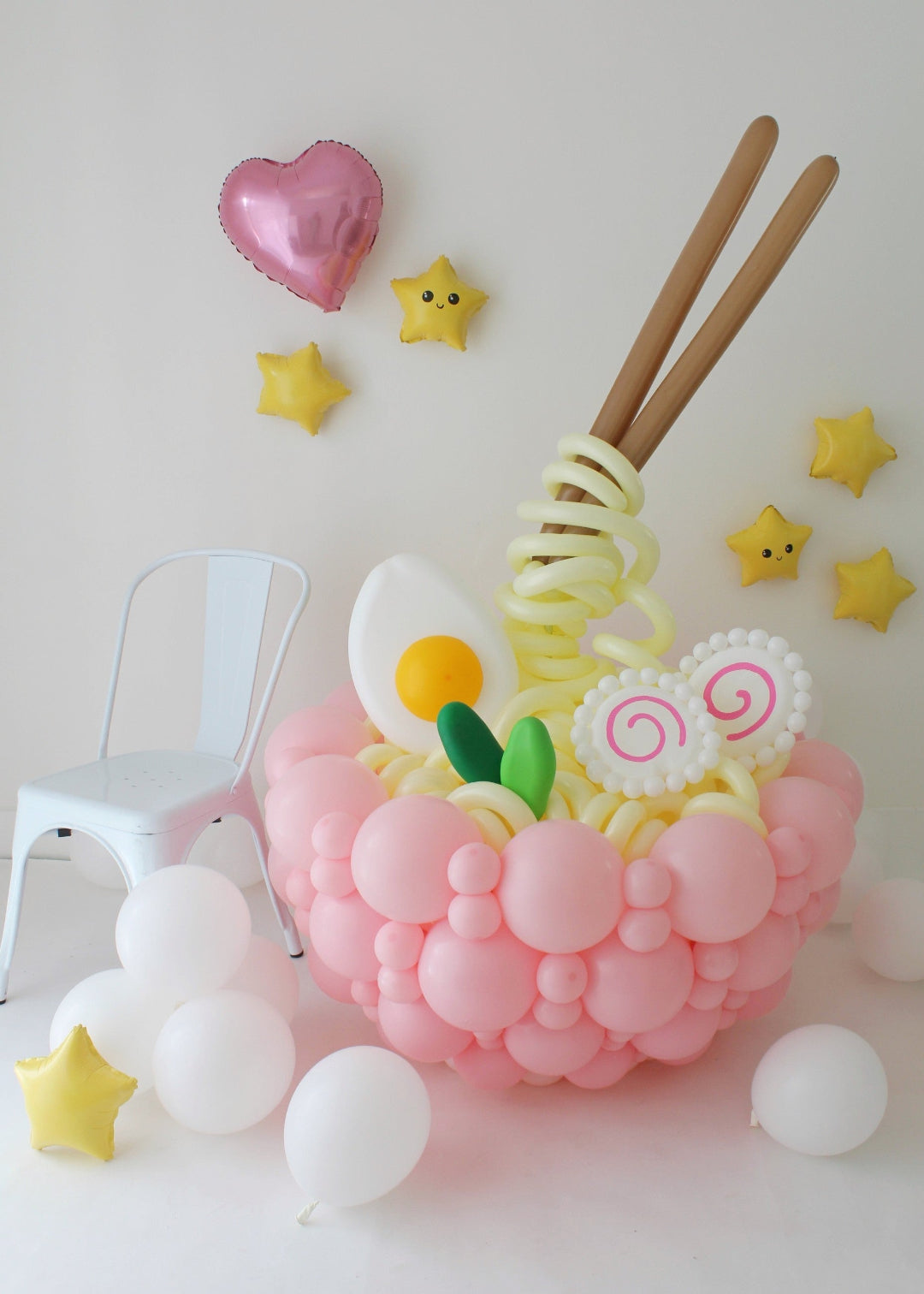 Balloon ramen bowl centerpiece with pink balloon base, spiral noodles, egg, fish cake, chopsticks, and star balloons on white background