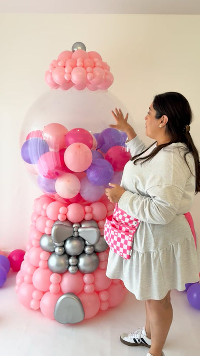 Lilly adjusting an oversized linking balloon gumball machine decor featuring a clear bobo globe filled with pink and purple mini balloons, a pink linking balloon base, and silver accent details. This behind-the-scenes image shows the scale and construction of a 7 to 7.5 foot tall freestanding gumball machine balloon design used for birthday parties, candy themed events, storefront displays, photo backdrops, and large scale balloon installs.