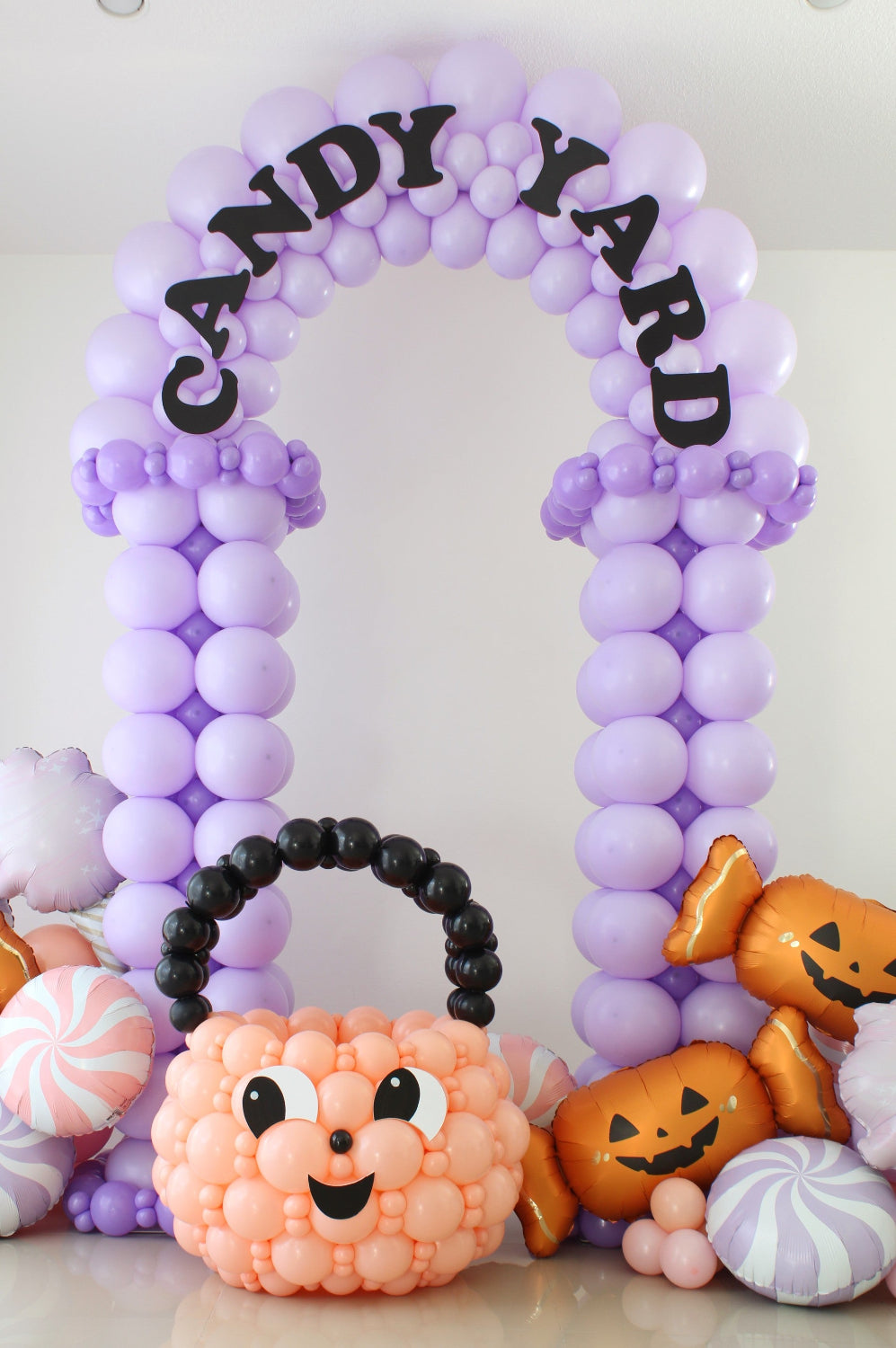 Lavender Candy Yard balloon arch with black lettering, a peach pumpkin balloon basket, and pastel candy and jack-o’-lantern foil balloons.