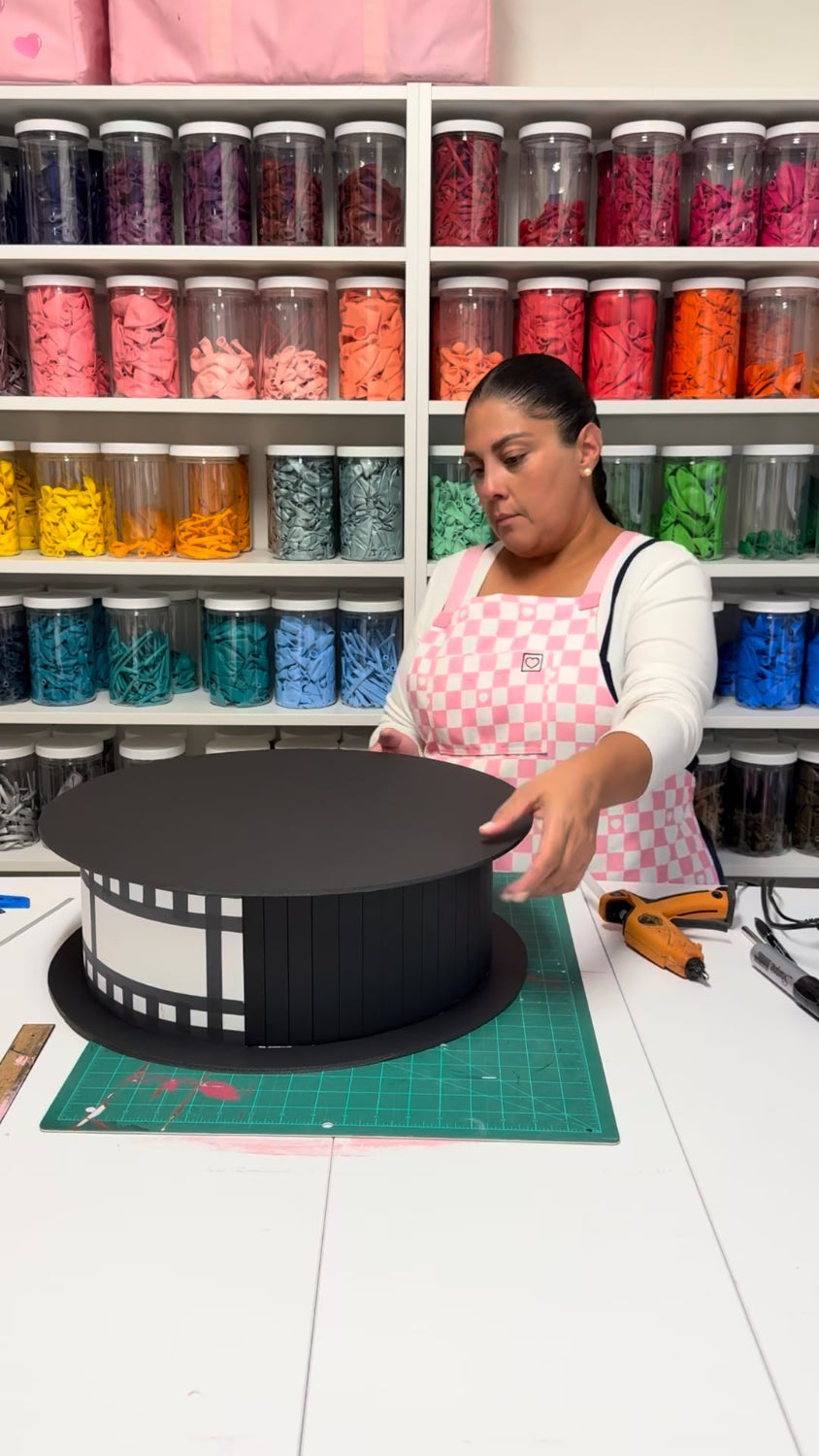 Lilly creating an oversized film reel prop with black foam board and film strip details for a cinema party decor class, movie night decorations, Hollywood themed events, and theater party displays.