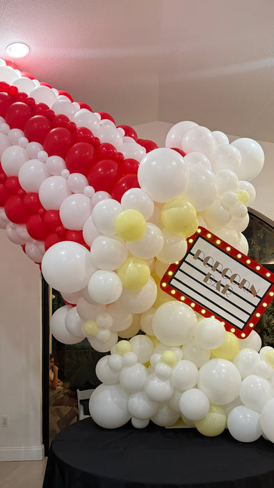 Giant balloon popcorn bucket close-up with red and white striped balloons, white popcorn balloon clusters, yellow butter accents, and a marquee popcorn sign for cinema party decor, movie night birthdays, Hollywood themed events, and watch party displays.