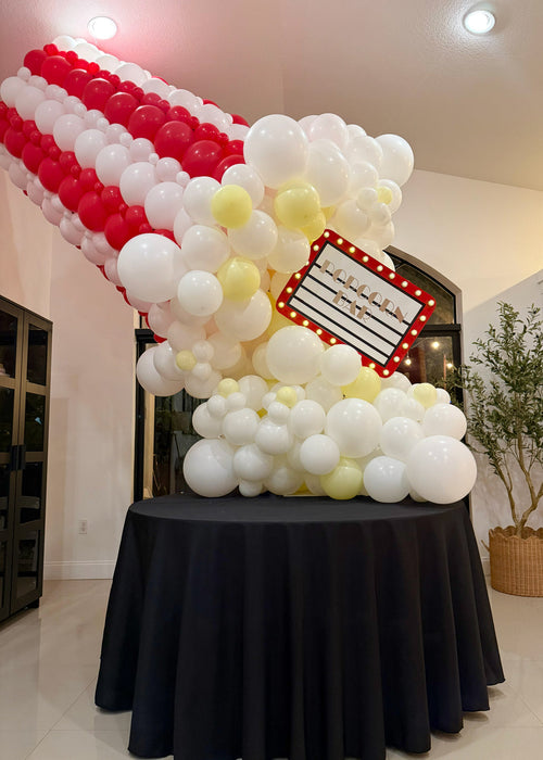 Giant balloon popcorn bucket made with red and white striped balloons, white popcorn balloon clusters, yellow butter accents, and a marquee popcorn sign for cinema party decor, movie night birthdays, Hollywood themed events, and watch party displays.