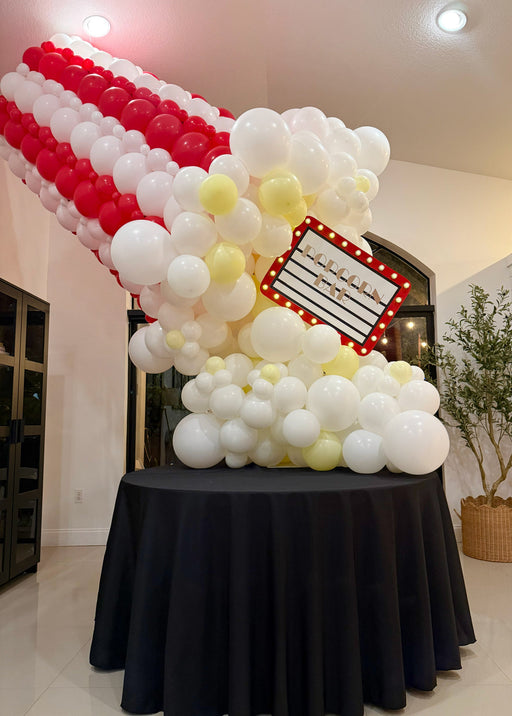 Giant balloon popcorn bucket made with red and white striped balloons, white popcorn balloon clusters, yellow butter accents, and a marquee popcorn sign for cinema party decor, movie night birthdays, Hollywood themed events, and watch party displays.