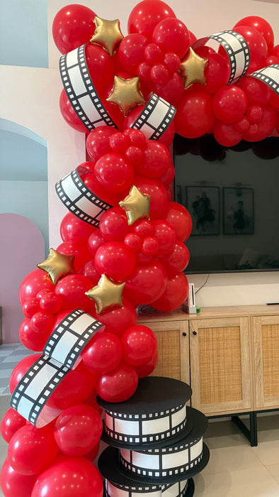 Red movie night balloon garland with film strip accents, gold star balloons, and stacked oversized film reel props for cinema party decor, Hollywood themed birthdays, award show events, theater parties, and watch party displays.