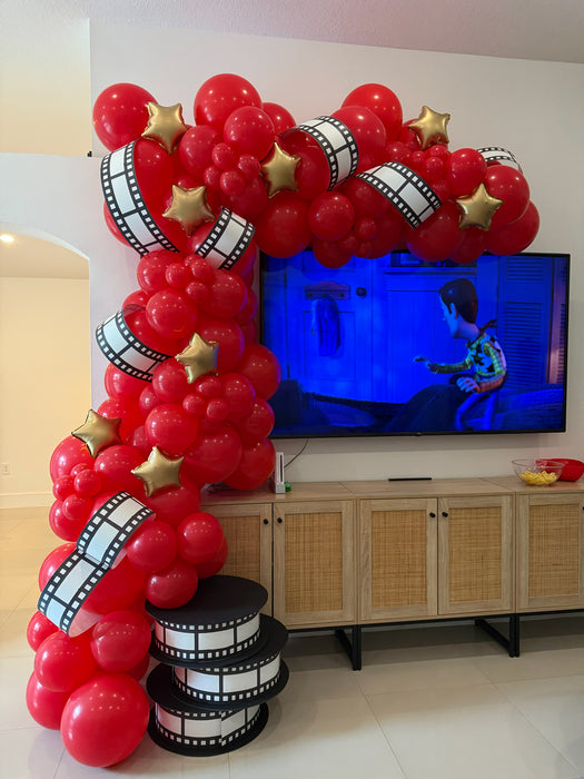 Red movie night balloon garland with film strip details, gold star balloons, and oversized film reel props for cinema party decor, Hollywood themed birthdays, award show events, theater parties, and family watch party displays.