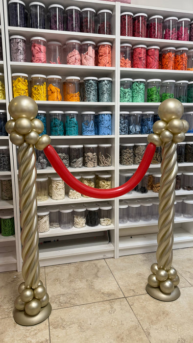 Red carpet balloon stanchions made with gold balloon columns and a red balloon rope for cinema party decor, movie night entrances, Hollywood themed parties, VIP areas, and photo op displays.