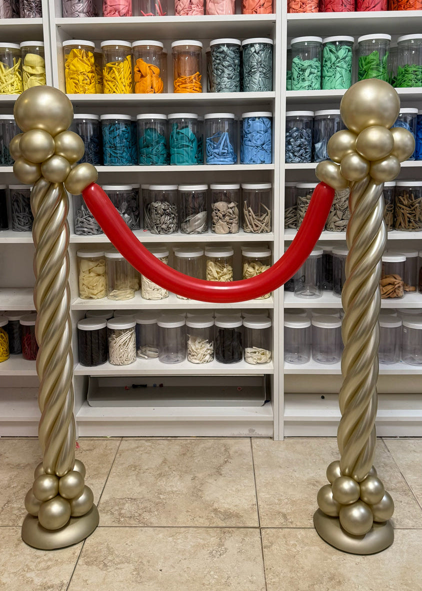 Gold balloon stanchions with a red balloon rope for red carpet entrances, cinema party decor, Hollywood themed birthdays, movie night events, award show parties, and VIP photo op displays.