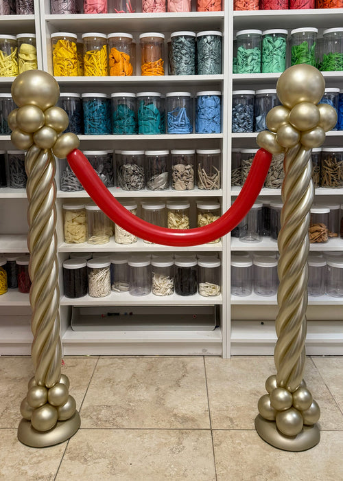 Gold balloon stanchions with a red balloon rope for red carpet entrances, cinema party decor, Hollywood themed birthdays, movie night events, award show parties, and VIP photo op displays.