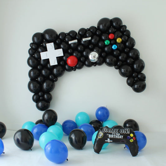 Oversized black game controller balloon sculpture with a white directional pad and colorful foil buttons above blue, aqua, and black party balloons.