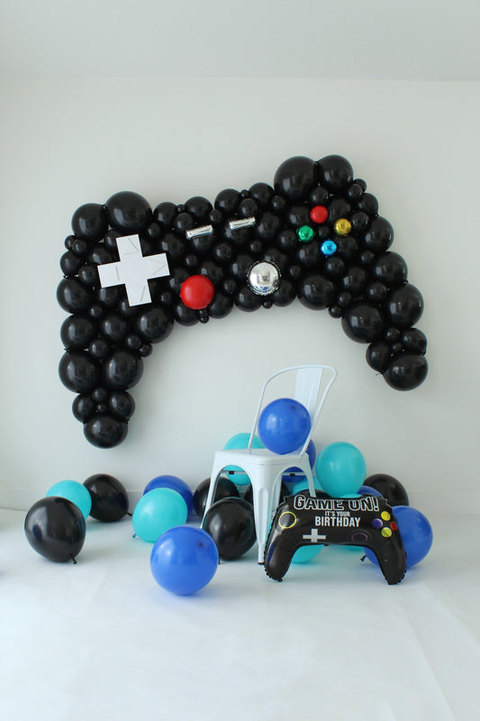 Oversized black balloon controller above a white chair, surrounded by blue, aqua, and black balloons and a “Game On!” birthday foil.