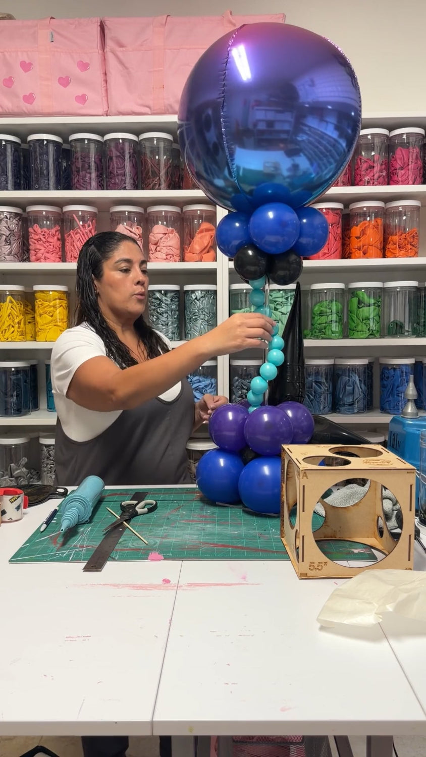Instructor assembling a gamer-themed balloon centerpiece with a blue-purple ombré orb, aqua balloon bubbles, and stacked blue and purple clusters.
