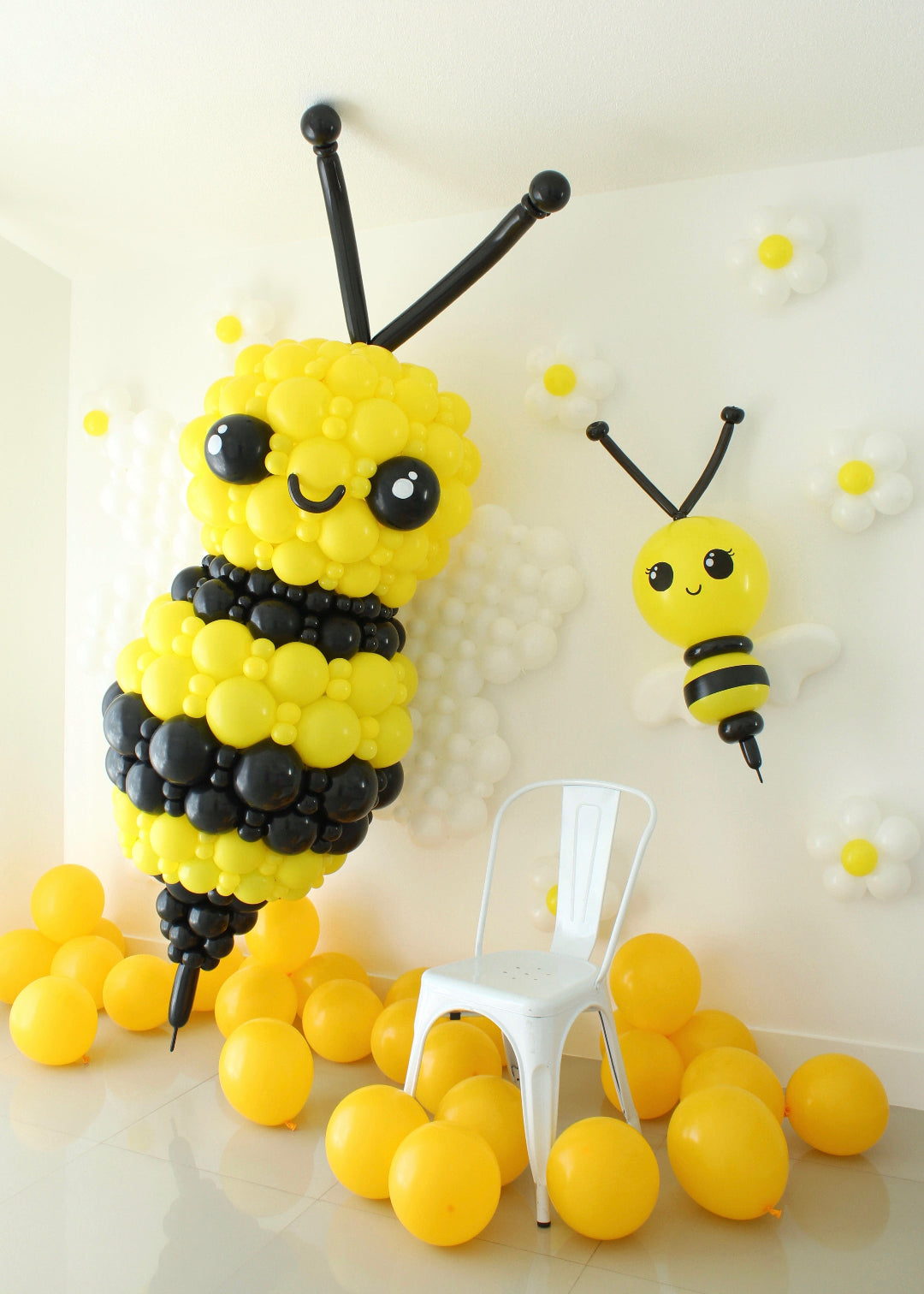 Giant balloon bumble bee sculpture created by Lilly Jimenez featuring layered yellow and black balloon clusters, oversized antennae and white wings. Large scale spring balloon decor displayed with yellow floor balloons and floral wall accents, ideal for garden theme parties, birthdays and professional event backdrops.