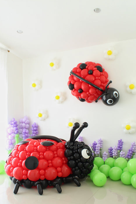 Large balloon ladybug sculpture with red clustered body and black detailing displayed in a spring garden balloon setup. Oversized bug decor paired with green balloon ground clusters and floral accents for birthdays and event backdrops.