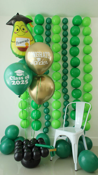 Graduation balloon bouquet featuring a Bravo-cado foil balloon, "Class of 2025" latex balloon, and personalized "Congrats Xander" gold balloon, anchored by a black graduation cap balloon weight. Displayed in front of a green balloon backdrop for a festive and fun graduation party setup.