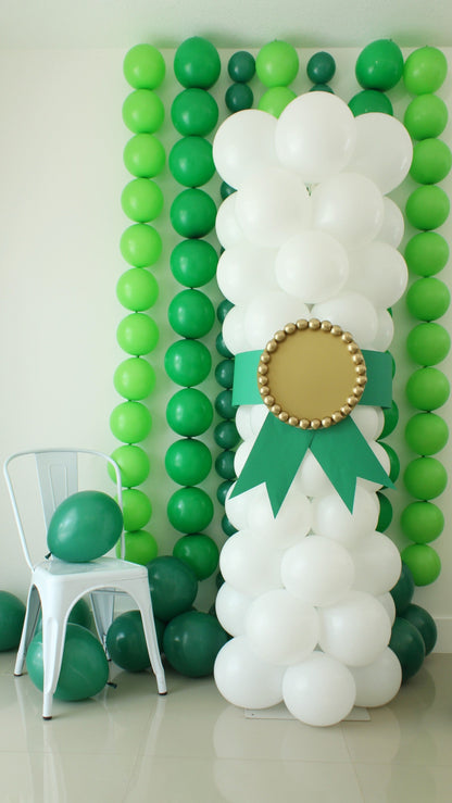DIY balloon column styled as a graduation diploma with white balloons, a green ribbon, and a gold medallion, placed in front of a green balloon wall backdrop with a white chair and green balloon accent. Ideal for graduation party decorations and photo setups.