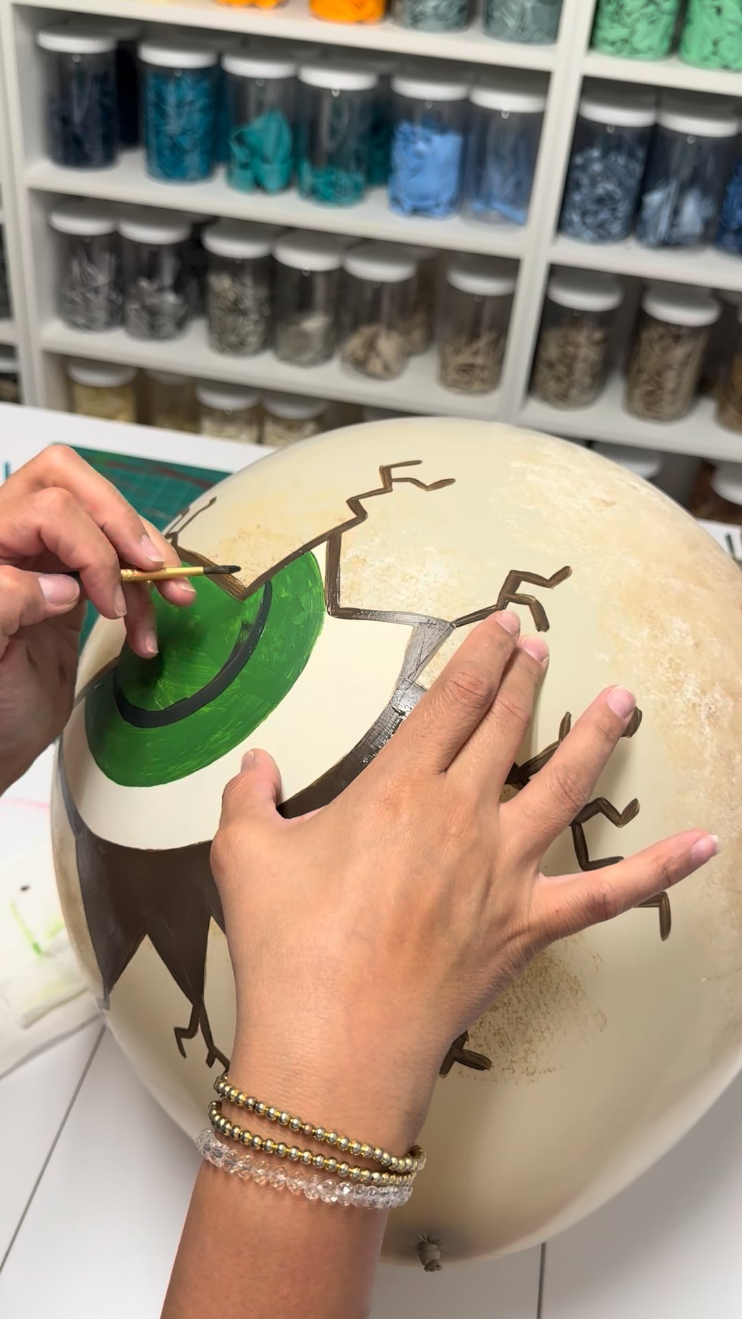 Close-up of hands painting a cracked dinosaur eye design on a beige balloon during a dinosaur egg balloon decor class.