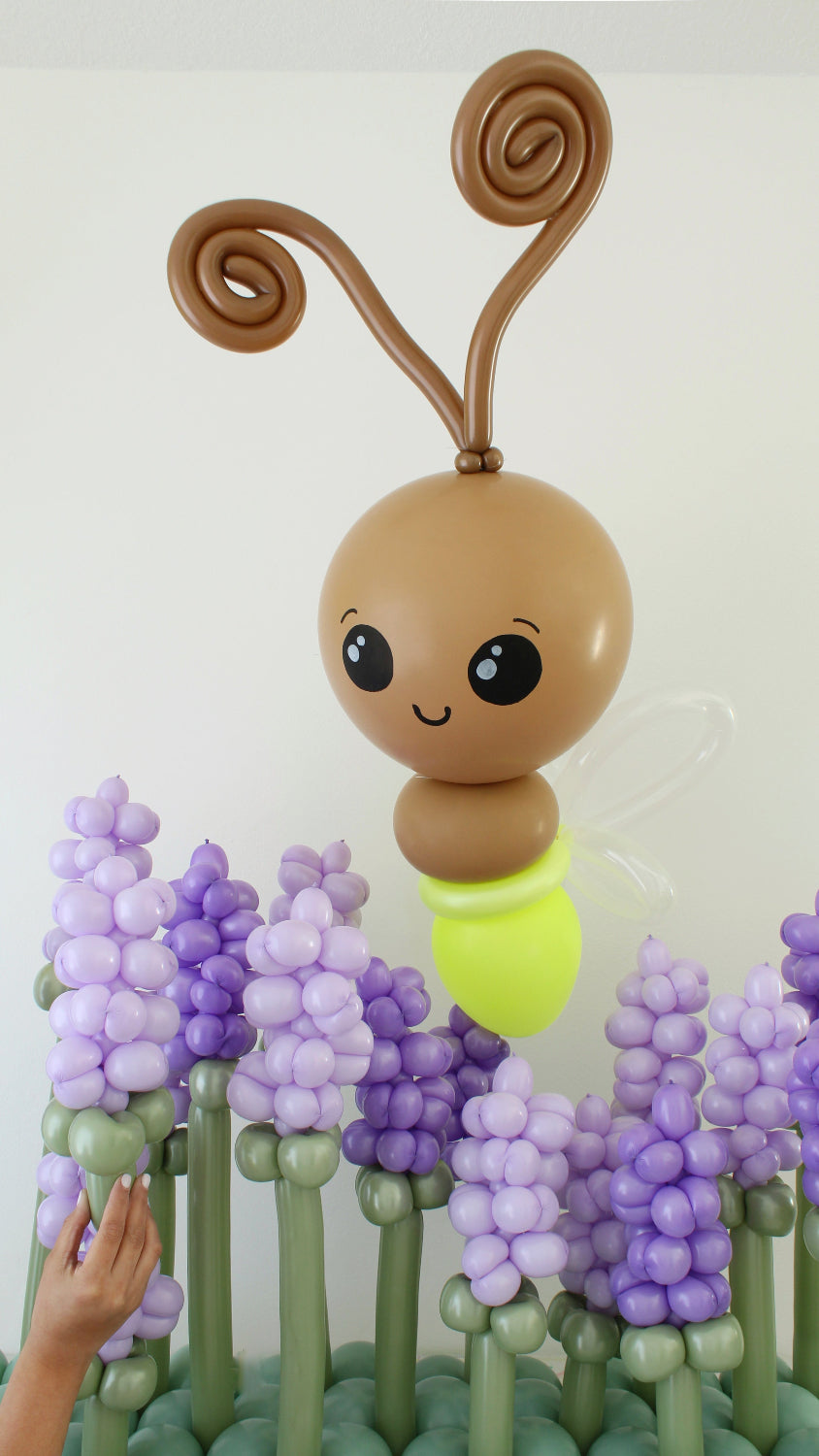 Giant light bug balloon character with spiral antennae and soft yellow body hovering above layered purple balloon flowers. Spring balloon decor design for garden themed parties, baby showers and professional balloon event styling.