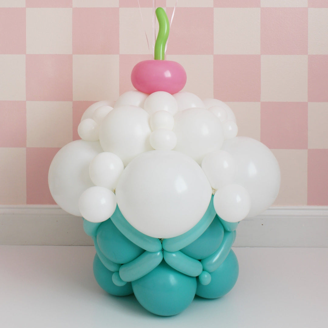 Balloon cupcake sculpture with white frosting, teal base, and cherry on top on pink checkered background