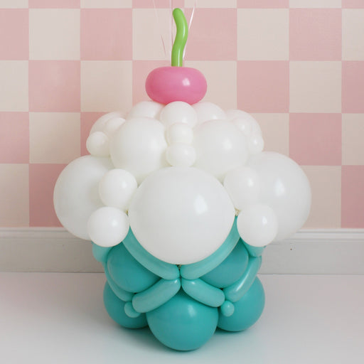 Balloon cupcake sculpture with white frosting, teal base, and cherry on top on pink checkered background
