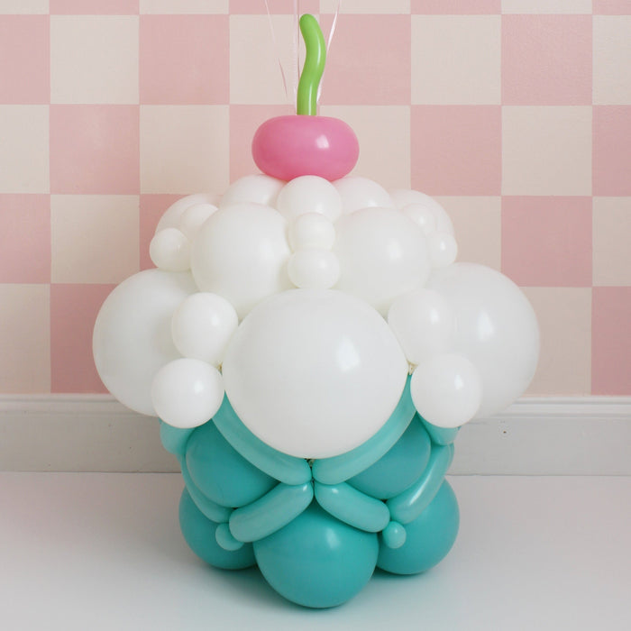 Balloon cupcake sculpture with white frosting, teal base, and cherry on top on pink checkered background