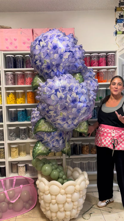 Balloon artist standing beside a large linking balloon flower pot topped with oversized blue hydrangea foil balloons and green balloon leaves.