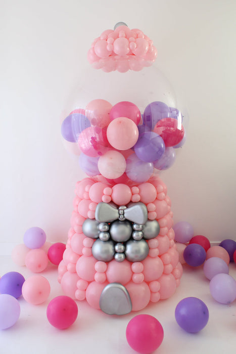 Oversized linking balloon gumball machine decor featuring a pink balloon base, silver accent details, and a clear bobo globe filled with pink and purple mini balloons. This freestanding balloon sculpture stands approximately 7 to 7.5 feet tall and is styled as a classic gumball machine for birthday parties, candy themed events, storefront displays, photo backdrops, and large scale balloon decor installs.