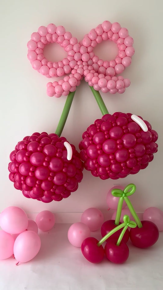 Oversized pink balloon cherries with a large light pink bow and green stems displayed with smaller balloon cherries.