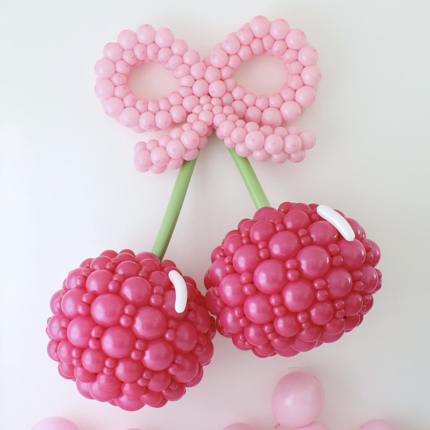 Oversized balloon cherry wall display featuring two hot pink cherries, green stems, and a large pink balloon bow.