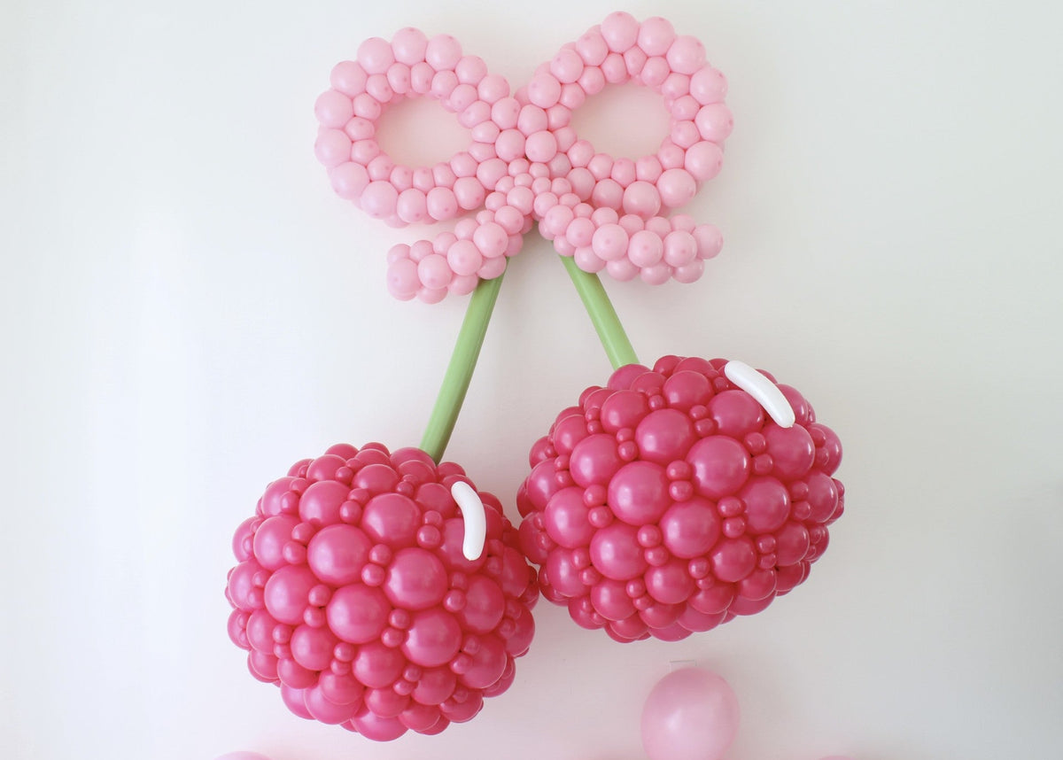Two oversized hot pink balloon cherries with green stems and a large light pink balloon bow displayed on a white background.