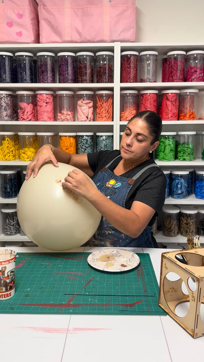 Balloon instructor hand-painting a beige balloon to create a realistic dinosaur egg in a colorful craft studio.