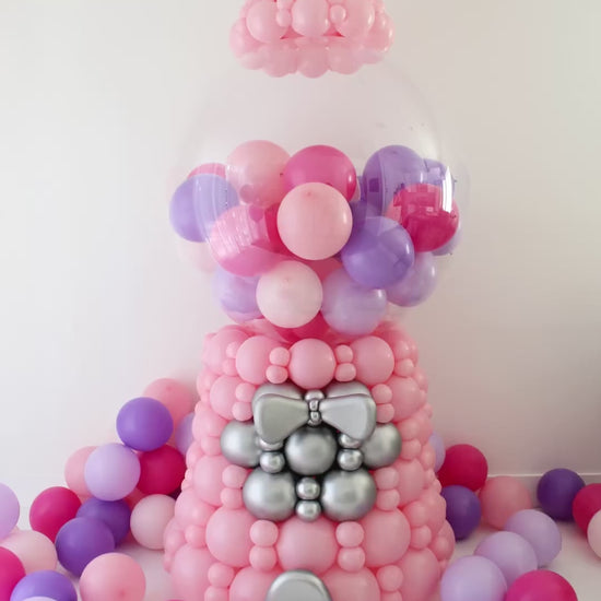 Animated view of an oversized linking balloon gumball machine decor featuring a pink balloon base, silver accent details, and a clear bobo globe filled with pink and purple mini balloons. The animation highlights the rotating gumball machine handle and front details, showing the scale and finished look of this 7 to 7.5 foot tall freestanding balloon design for birthday parties, candy themed events, storefront displays, photo backdrops, and large scale balloon installs.