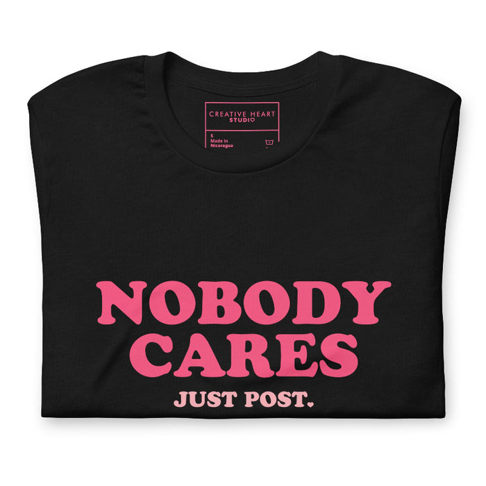 folded black t-shirt featuring the phrase "Nobody Cares, Just Post" in bold pink and white lettering, perfect for content creators and casual wear.