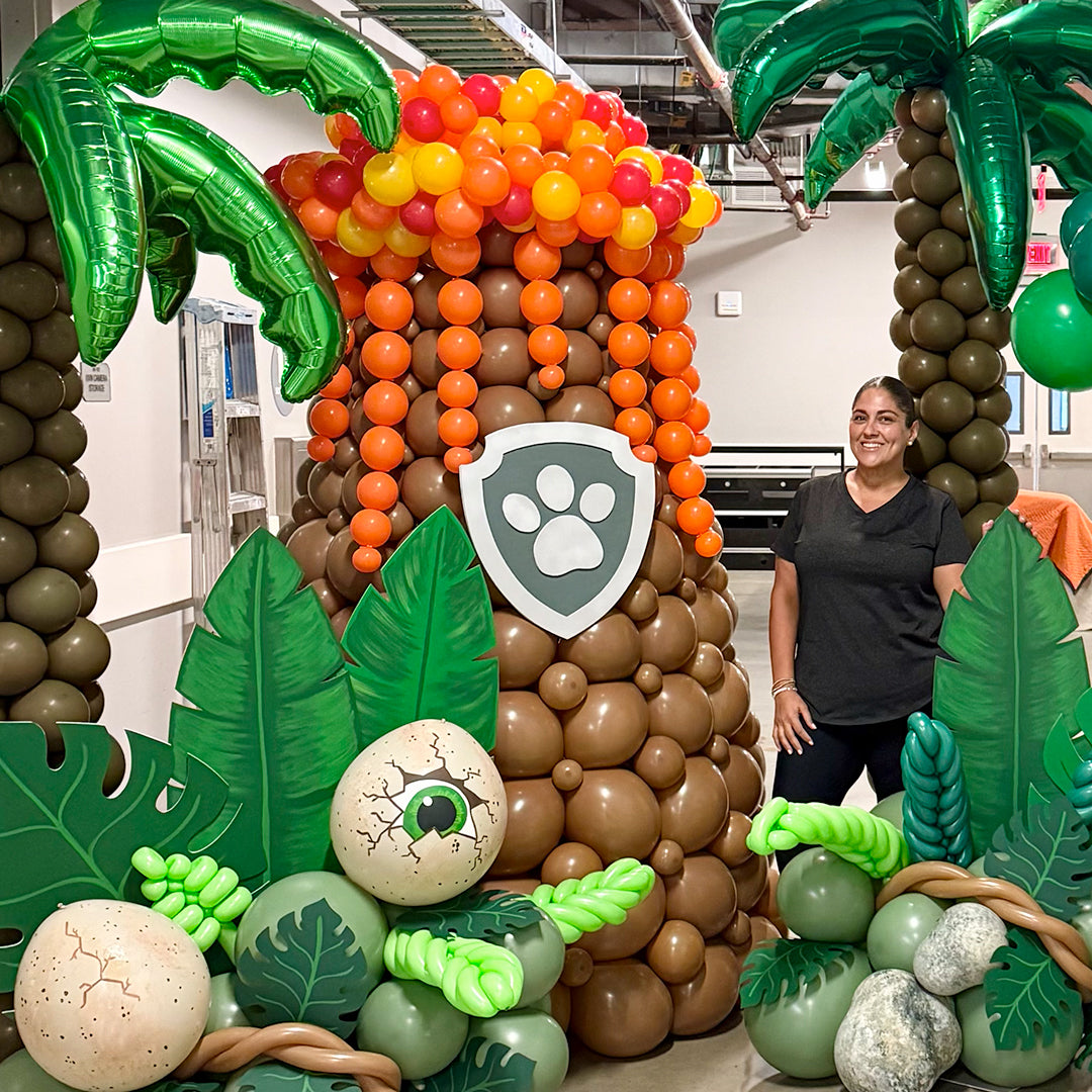 Large balloon volcano with orange lava, tropical palm trees, jungle leaves, dinosaur eggs, and themed party props for a prehistoric event display.