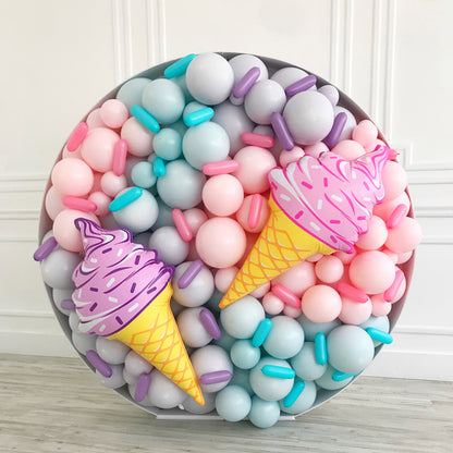 A circular ice cream-themed balloon mosaic wall featuring pastel balloons in pink, lavender, gray, and teal. Two oversized ice cream cone cutouts with colorful sprinkle details sit on top of the balloon base, creating a sweet and whimsical display. Designed by Lilly Jimenez, this piece is part of the Balloon Mosaic® Walls Course—perfect for summer parties, dessert tables, or Instagram-worthy backdrops.