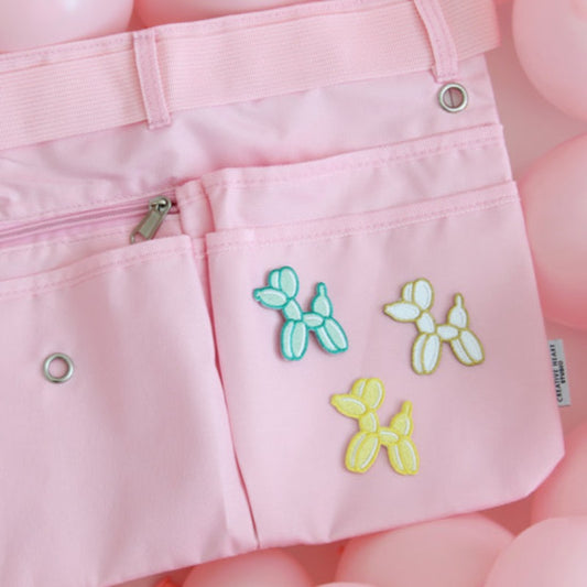 Light pink maker apron featuring three embroidered balloon dog patches in teal, white, and yellow on the front pocket. The apron includes multiple pockets, a center zip pouch, belt loops, and grommets for tool attachments. Displayed on a background of pink balloons, this stylish and functional workbelt is perfect for balloon artists, event decorators, and creative professionals looking to add a playful and personalized touch to their gear.