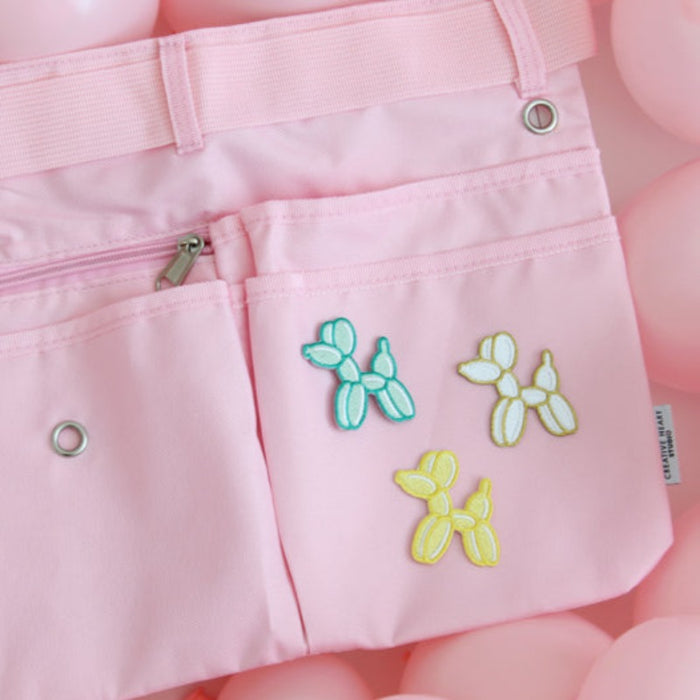 Light pink maker apron featuring three embroidered balloon dog patches in teal, white, and yellow on the front pocket. The apron includes multiple pockets, a center zip pouch, belt loops, and grommets for tool attachments. Displayed on a background of pink balloons, this stylish and functional workbelt is perfect for balloon artists, event decorators, and creative professionals looking to add a playful and personalized touch to their gear.